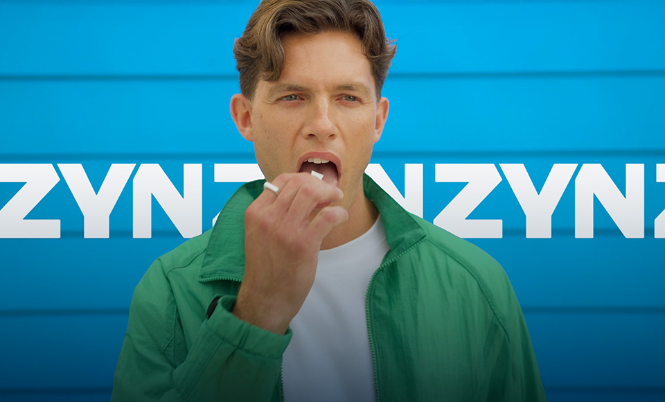 Man wearing a white shirt and green sweatshirt trying a ZYN nicotine pouch in front of a ZYN logo